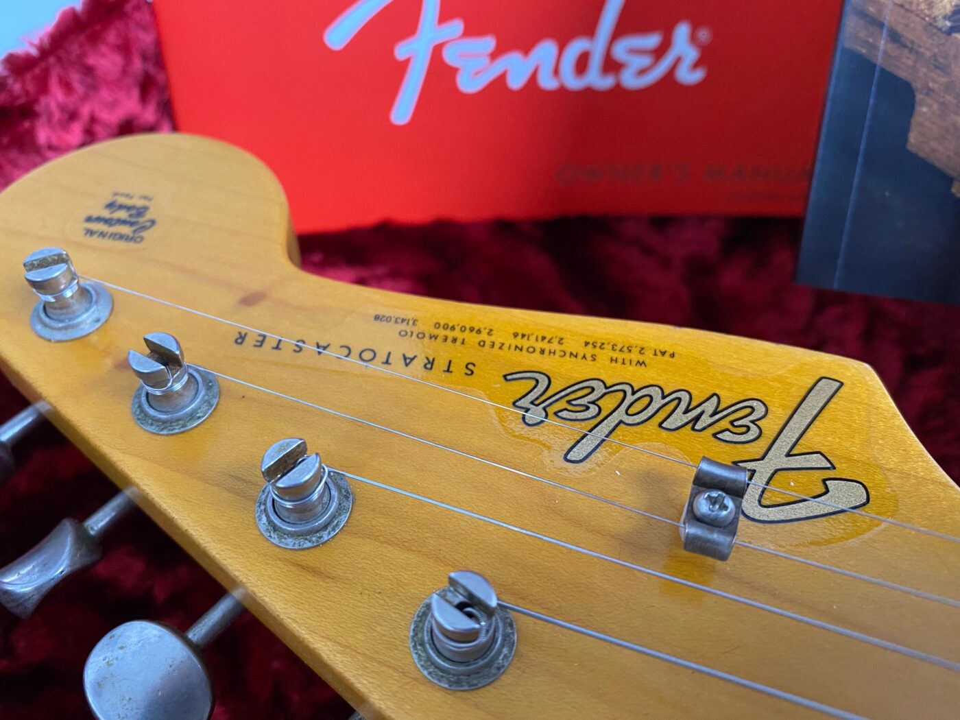 Close-up of a Fender Stratocaster guitar headstock with tuning pegs, showcasing the Fender logo and string details on a red velvet background.